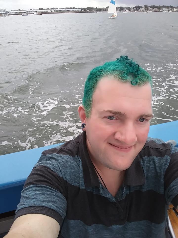 Image of man with blue hair taking selfie on boat with water behind him. 